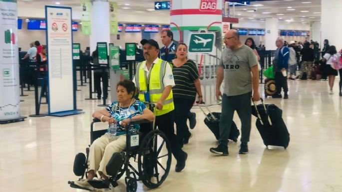 A person in a wheelchair being assisted by an airport employee, with other travelers walking around in the airport terminal.