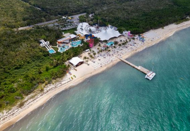 Aerial image showing a beach resort with buildings and a pier extending into clear blue waters, surrounded by trees and a sandy beach.