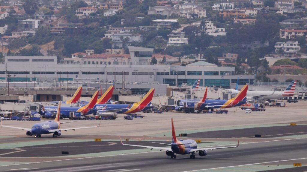 Multiple airplanes on the ground at an airport with airport infrastructure in the foreground and hills with houses in the background.