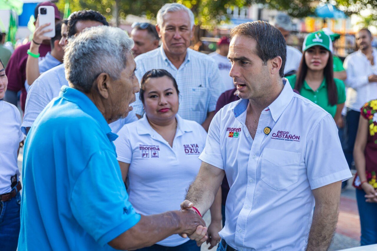 An older man in a blue shirt shaking hands with a smiling man in a white shirt with political campaign logos, surrounded by onlookers.