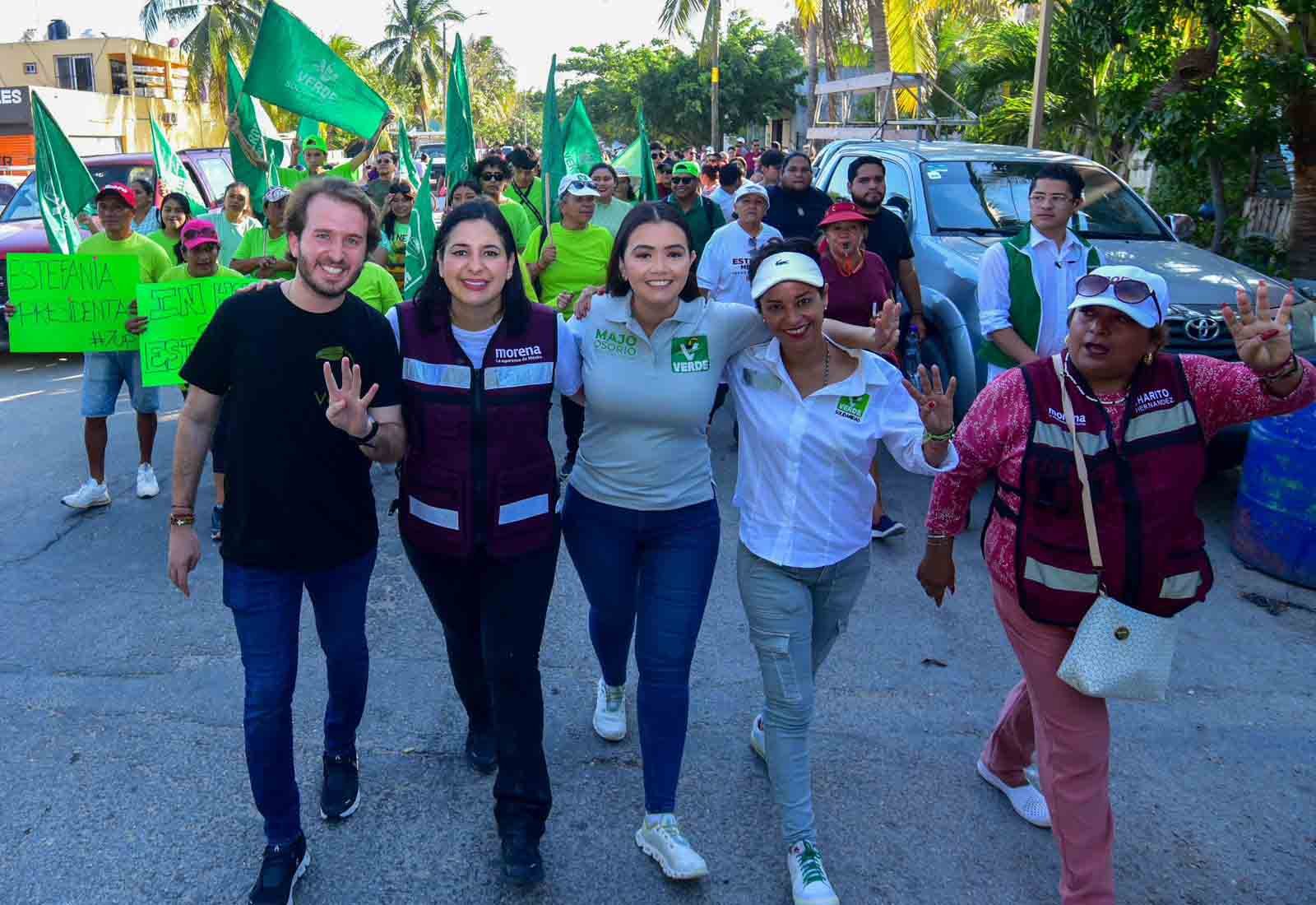 A group of individuals participating in a political rally, with some waving to the camera and others holding banners and green flags, conveying a sense of community activism and engagement.