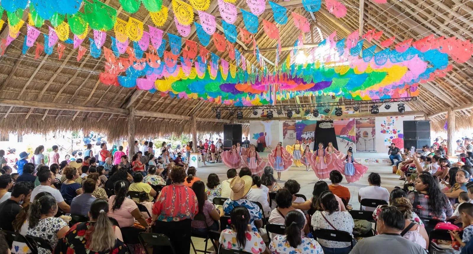 People attending a dance performance with performers in traditional attire under colorful paper decorations in an open-air pavilion.