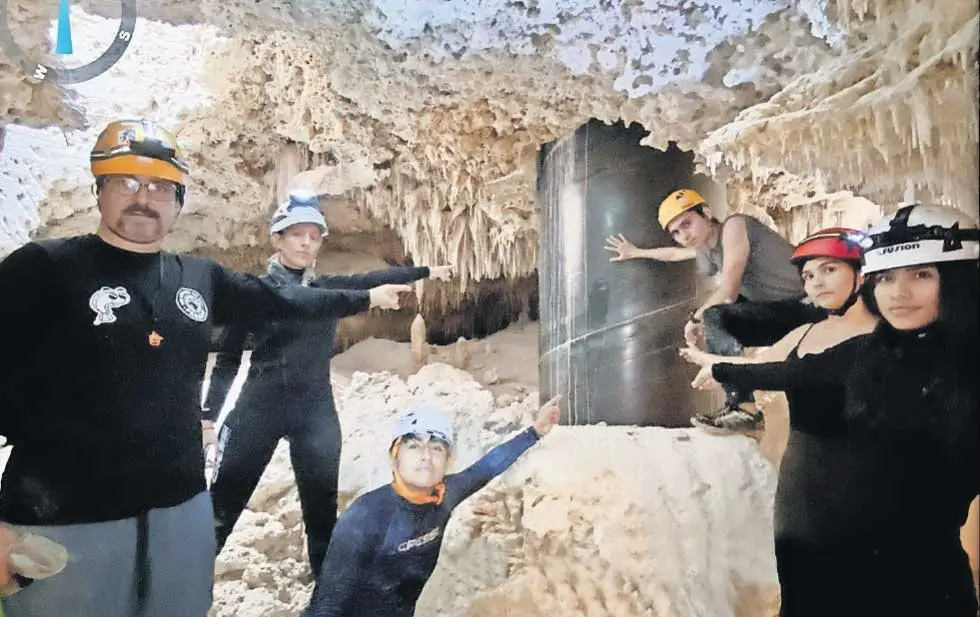A group of six explorers wearing helmets and headlamps in a cave with stalactites and stalagmites, pointing at and admiring a reflective surface on the cave wall.