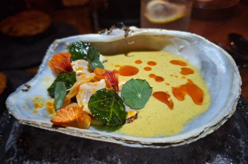 An elegantly presented seafood dish in a large half shell, garnished with herbs and edible flowers, with a creamy sauce and drizzle of orange oil.