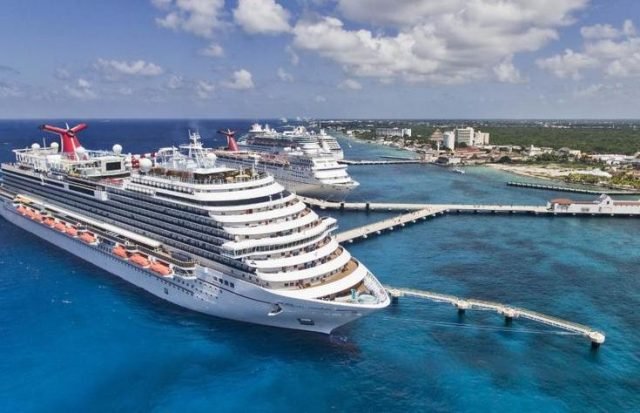 Aerial view of two large cruise ships docked at a tropical port with clear blue waters and coastal buildings in the background.