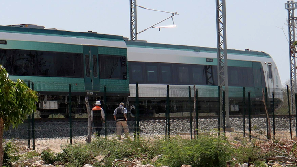 Two individuals wearing hard hats observe a green and white train passing on railway tracks surrounded by a fenced area with sparse vegetation and debris on the ground.