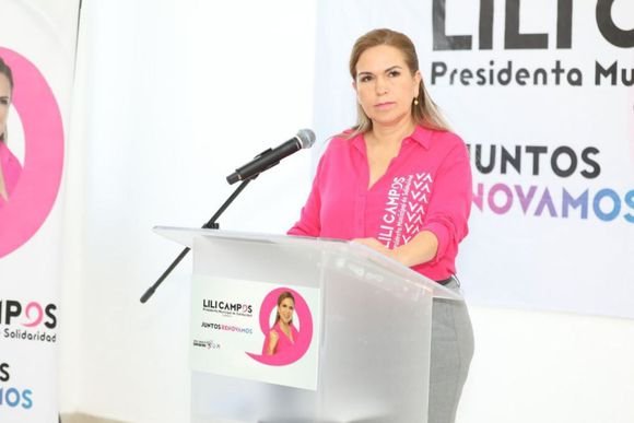 A woman in a pink shirt standing at a podium with microphones, with banners displaying her name and slogans in the background
