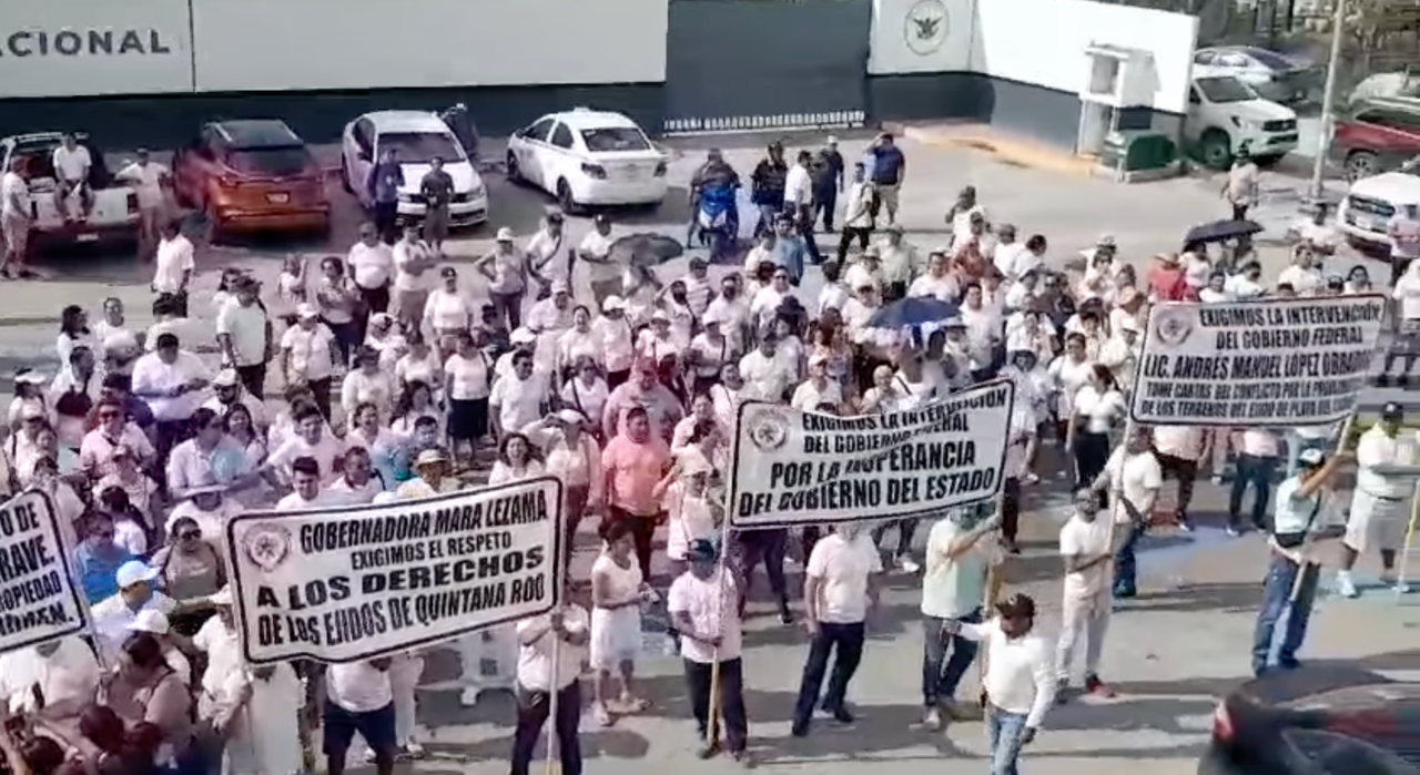 A group of protestors in white shirts carrying banners with text demanding respect for land rights and government intervention in Quintana Roo, Mexico.