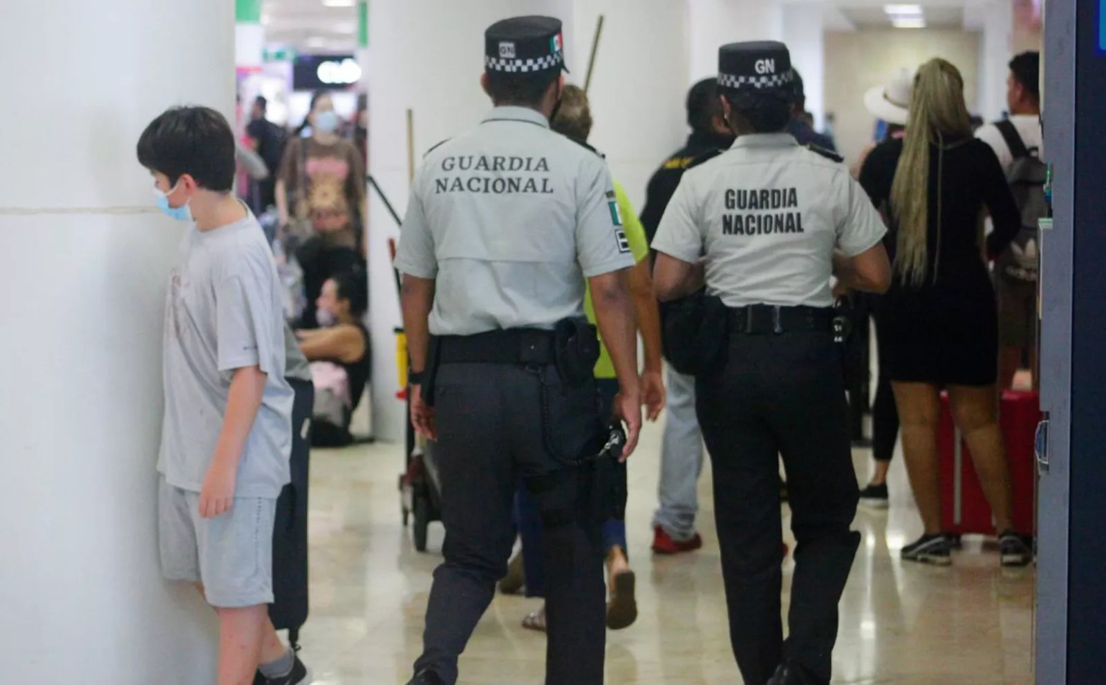 Two uniformed Guardia Nacional officers patrolling a crowded corridor with civilians, including a person wearing a face mask looking in their direction.