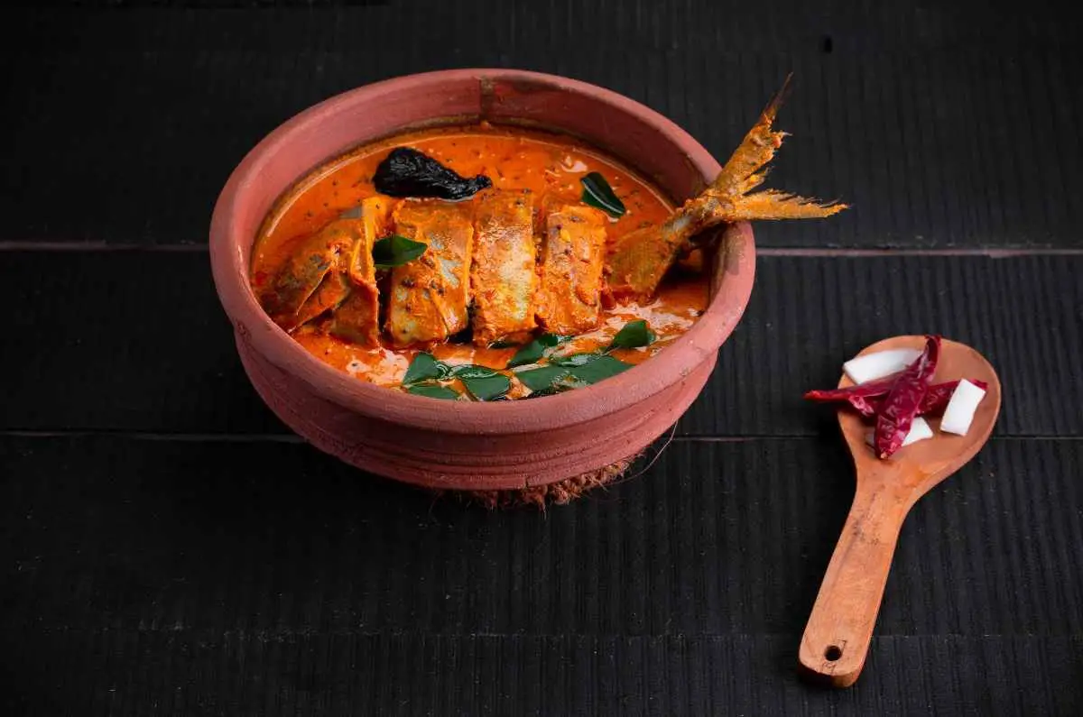 A clay pot filled with fish curry, garnished with curry leaves, accompanied by a wooden spoon with chillies and coconut pieces on a dark surface.