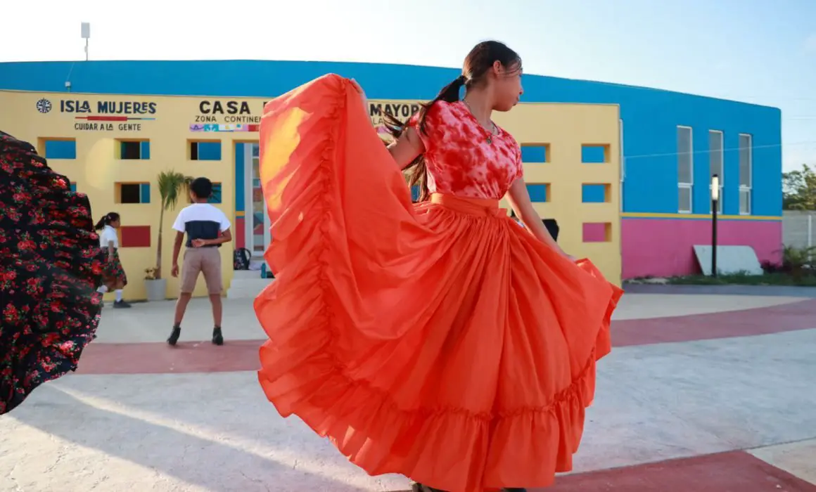 A woman twirls in a bright orange traditional dress with flowing skirt in front of a colorful building, while people walk by in the background