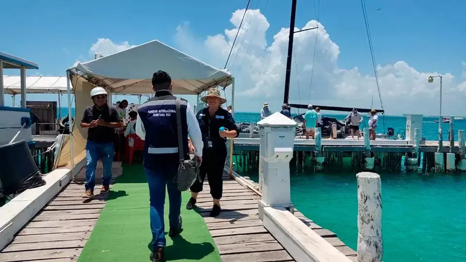 People on a vibrant dock with a clear blue sky and turquoise ocean in the background. Some individuals appear to be engaging in conversation or heading towards boats moored alongside the wooden pier. A canopy provides shade, while a worker in a vest walks towards the viewer.