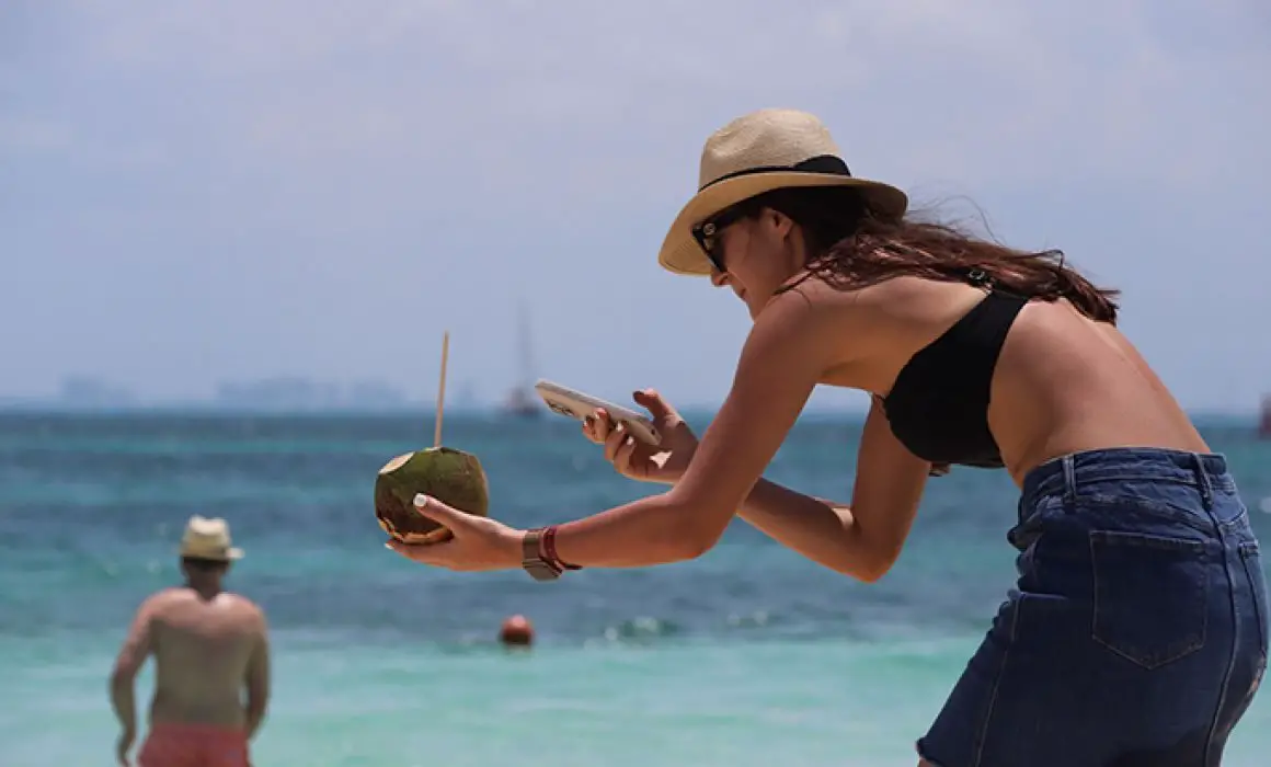 A woman in a sunhat and sunglasses crouches to photograph a held coconut with her smartphone at a sunny beach, with the ocean and a standing man in the background.