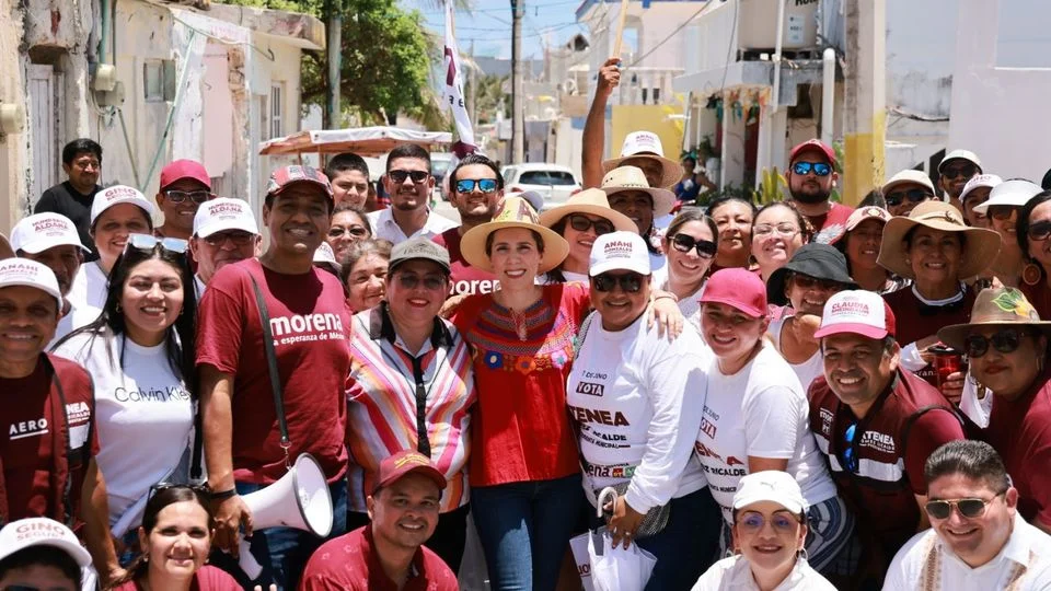 A group of smiling people wearing red and white hats and shirts, some with the word 'morena' printed on them, gathered on a sunny street