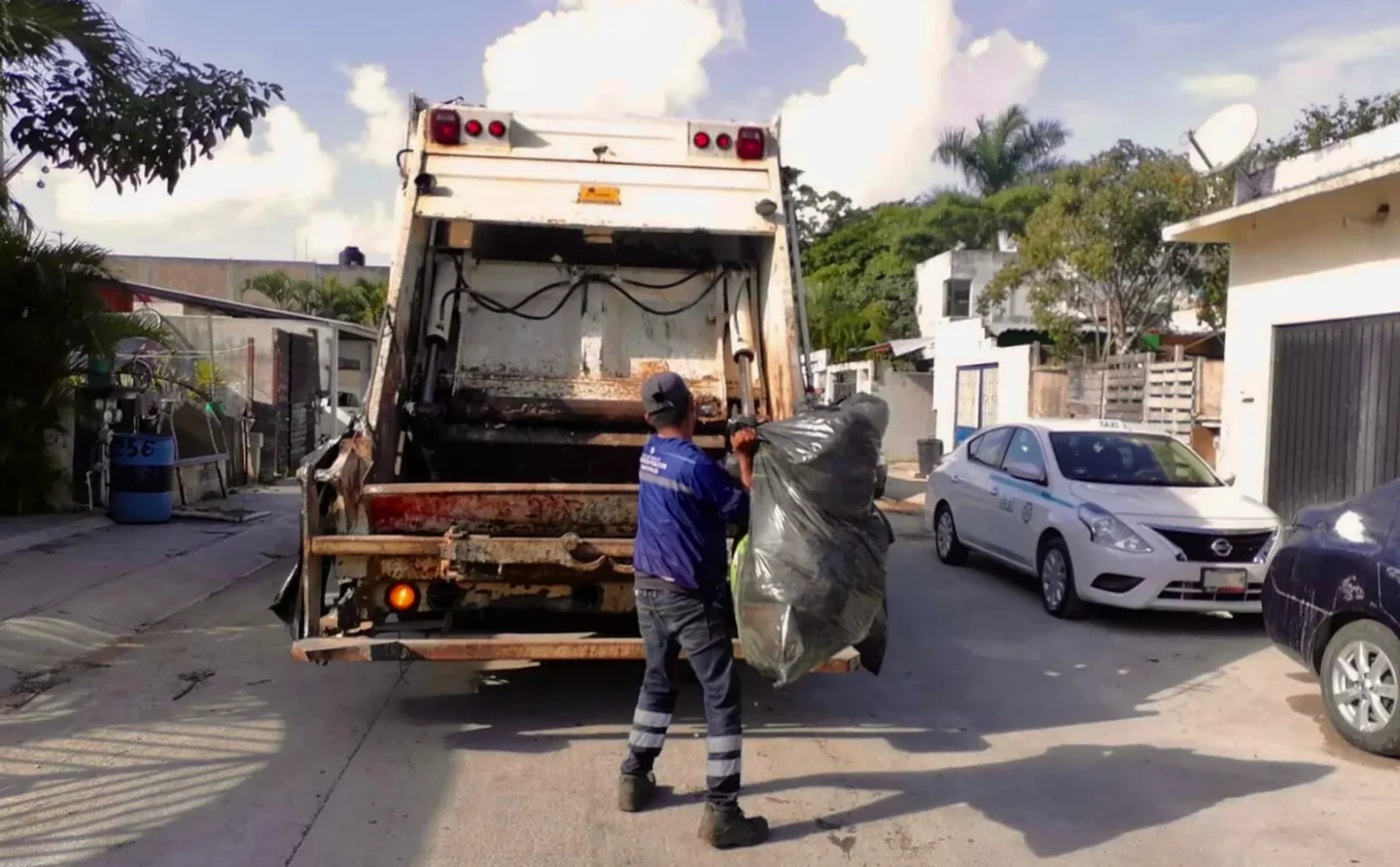 A worker in a blue uniform lifting a large trash bag into a garbage truck on a sunny residential street.