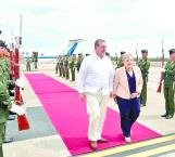 Two individuals walking on a red carpet with honor guards lined up on the side against a backdrop of an airplane and overcast sky