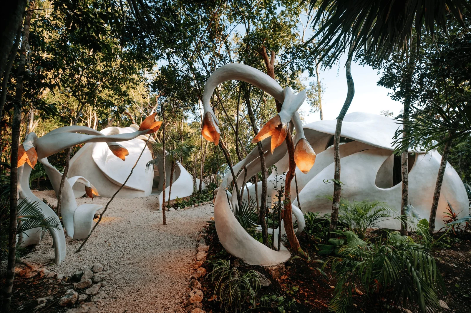 Unique sculptural white buildings with organic shapes nestled among tropical forest vegetation, giving the impression of futuristic habitation in a natural setting. Pathways are illuminated by warm-hued lanterns amid the greenery.