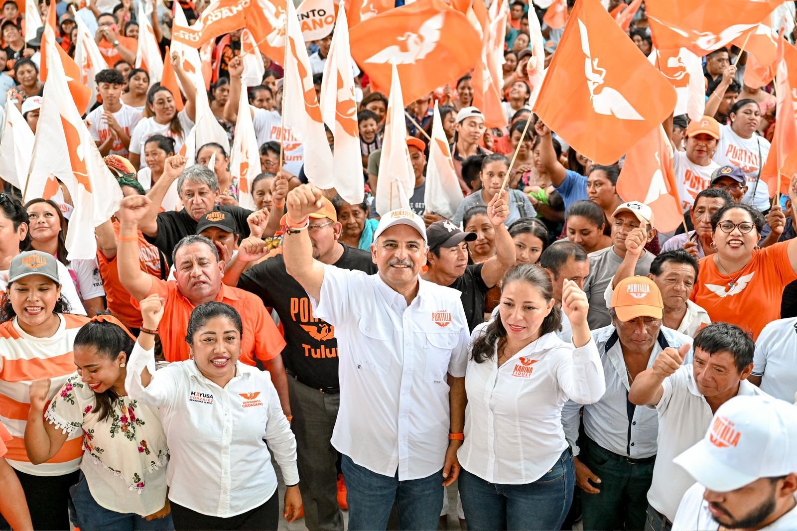 A group of people at a political rally waving orange flags and showing support with raised fists, some wearing branded clothing and hats.