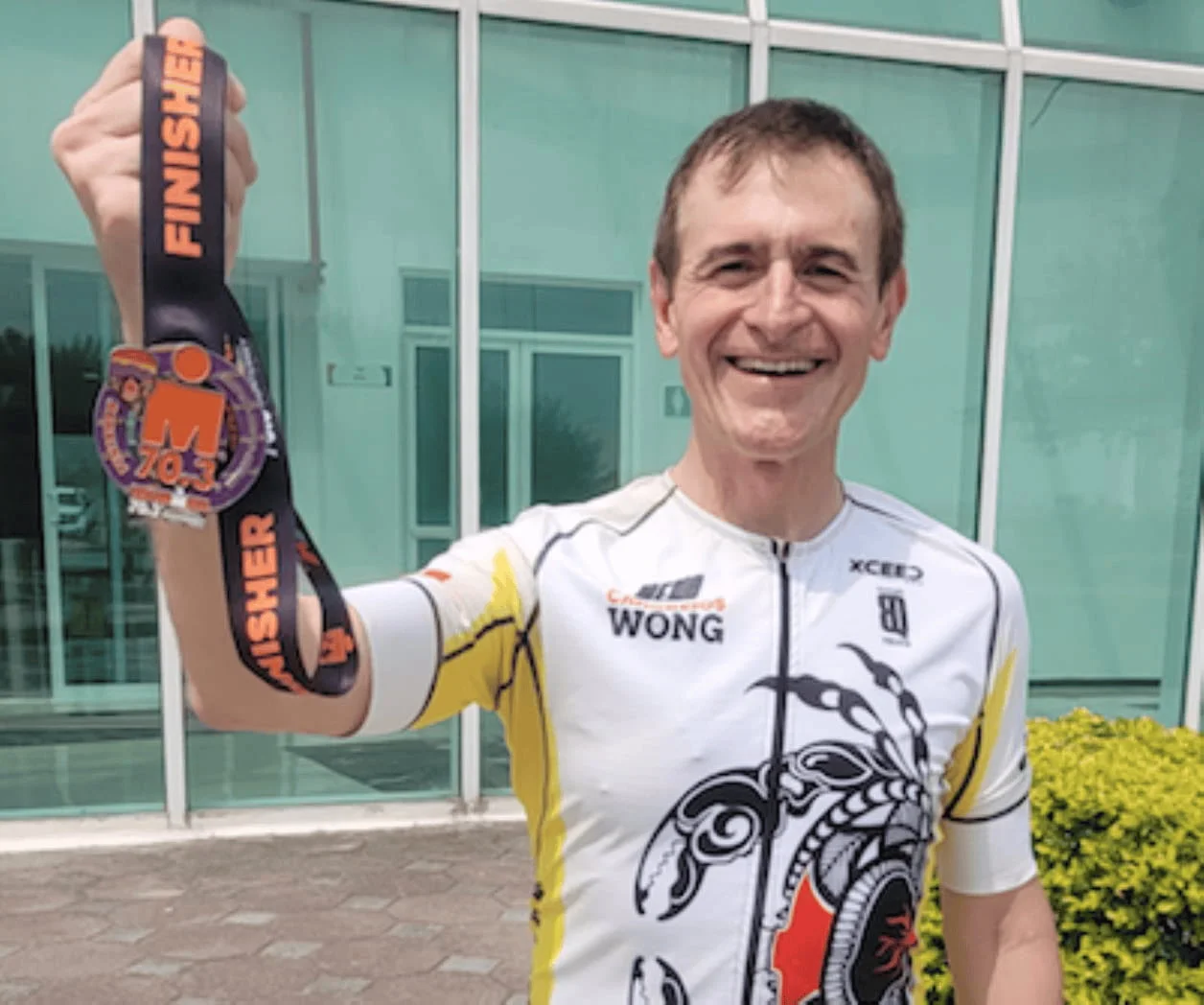 A smiling person in a cycling jersey holding up a 'Finisher' medal outside a building with glass doors.