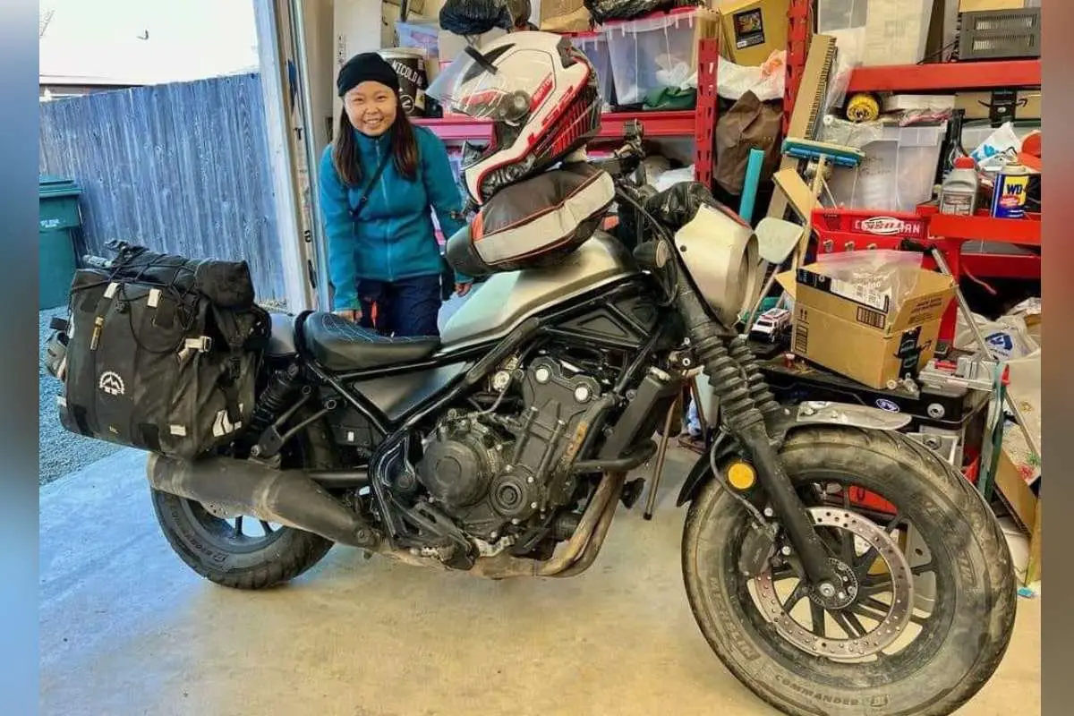 A person standing smiling next to a black motorcycle equipped with luggage, inside a garage full of various items.