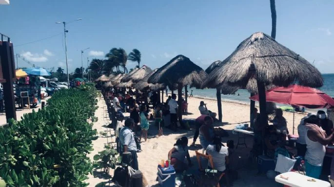 People enjoying a sunny day at a beach market with thatched umbrellas along the shore and various stalls and seating areas.