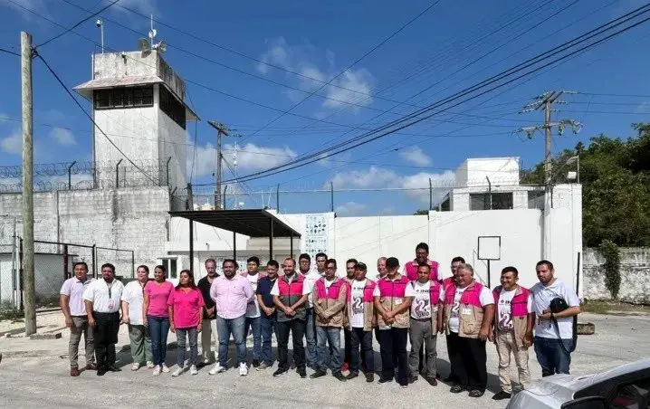 A group of people posing for a photograph in front of a correctional facility with barbed wire and a watchtower in the background
