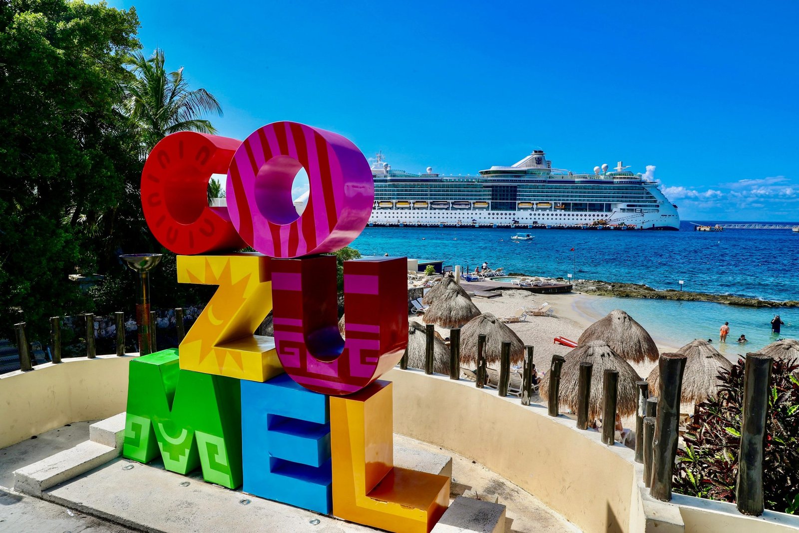 Colorful "COZUMEL" sign in the foreground with a cruise ship and beach-goers in the background on a sunny day