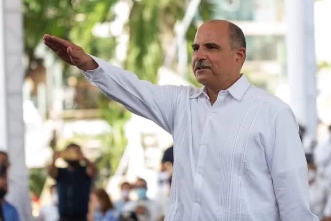 A man in a white shirt gesturing while speaking to an audience outside with trees and people in the background.