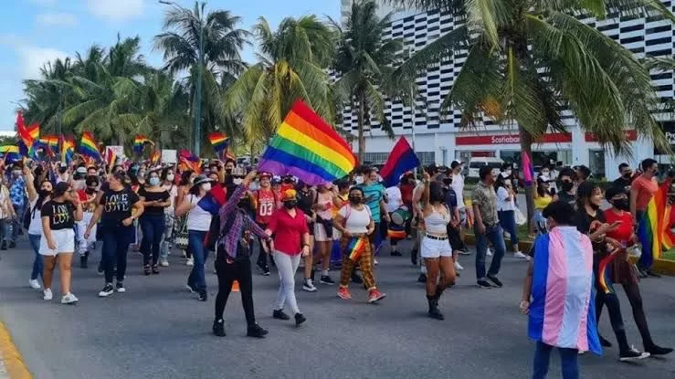 A lively pride parade with participants holding rainbow flags and marching on a street lined with palm trees and buildings.