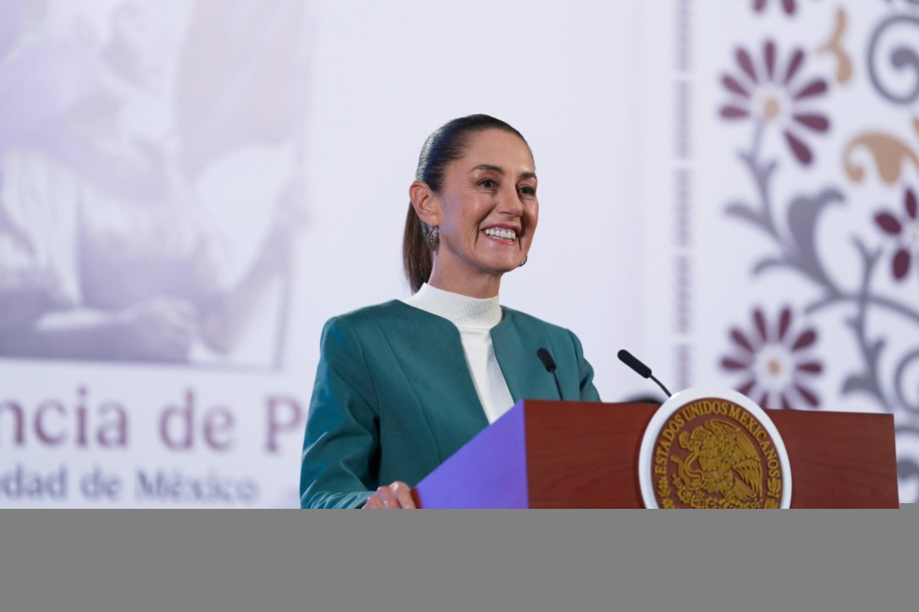 A smiling woman in a green blazer speaking at a lectern with a microphone and a national emblem.