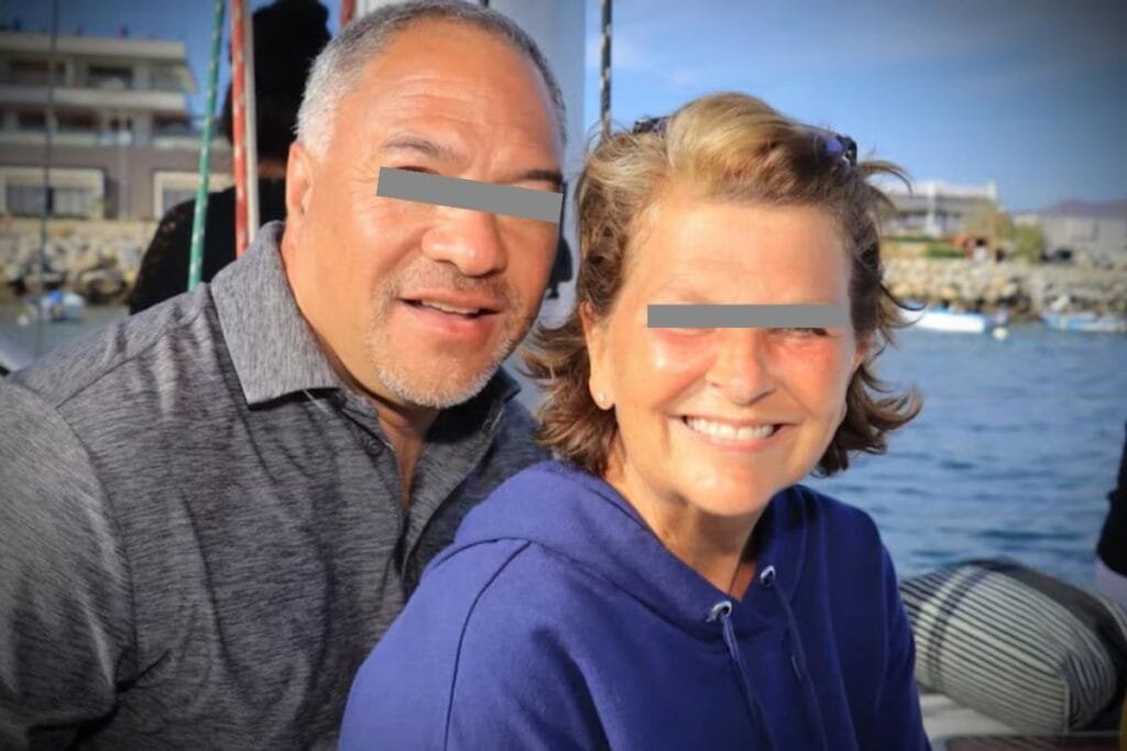 Two smiling adults with obscured eyes, one male and one female, by the seaside with a harbor in the background.