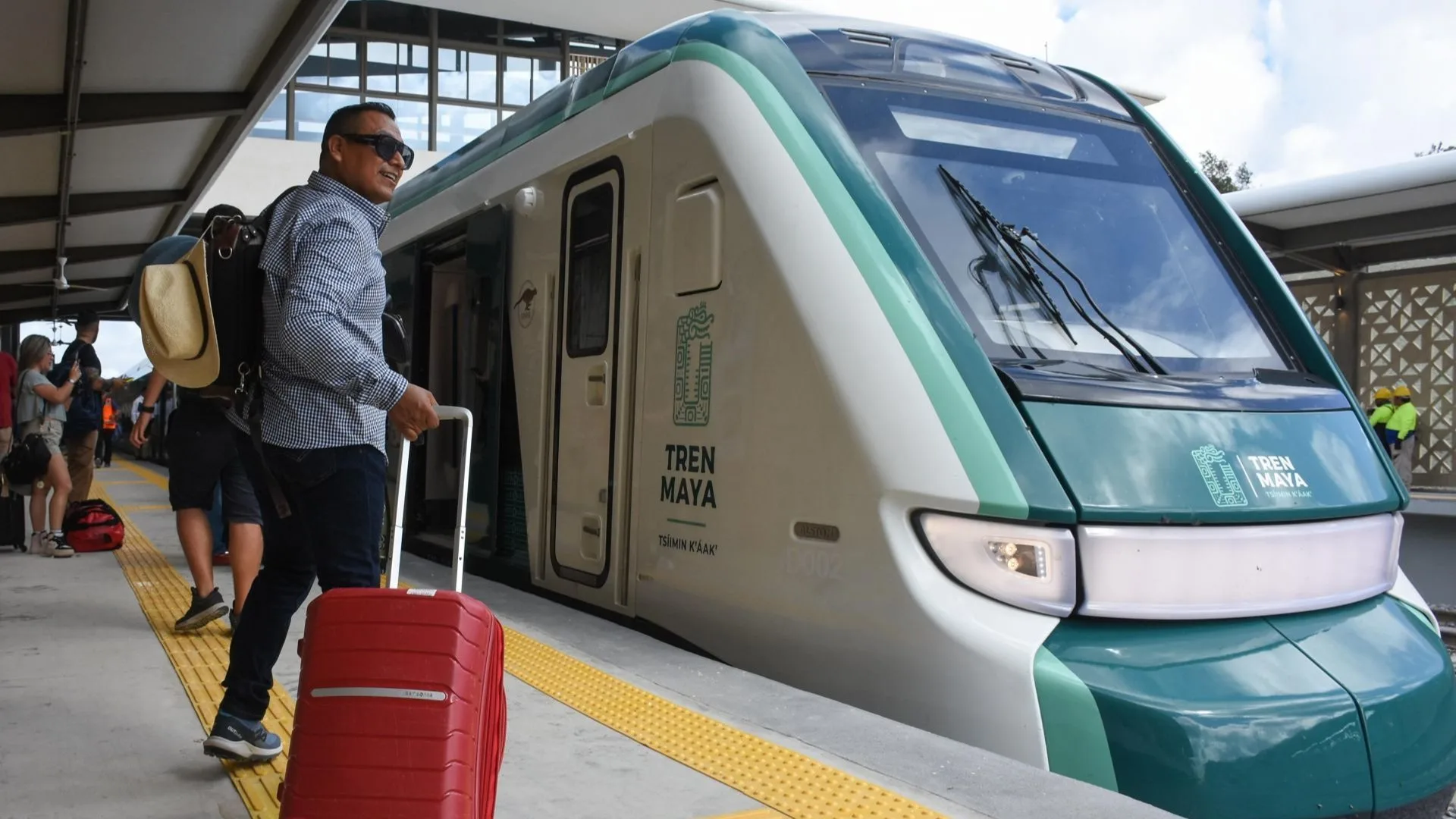 A person with a red suitcase prepares to board the Tren Maya train at a station, wearing sunglasses and carrying a bag on their shoulder. Other passengers can be seen in the background.$# CAPTION