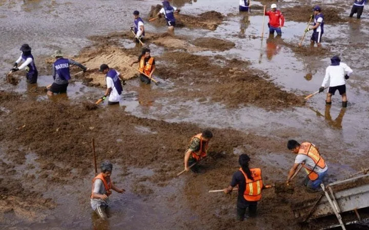 Volunteers working together to clean a muddy wetland area, with some wading in water and others on the dry patches, under a clear sky.$# CAPTION