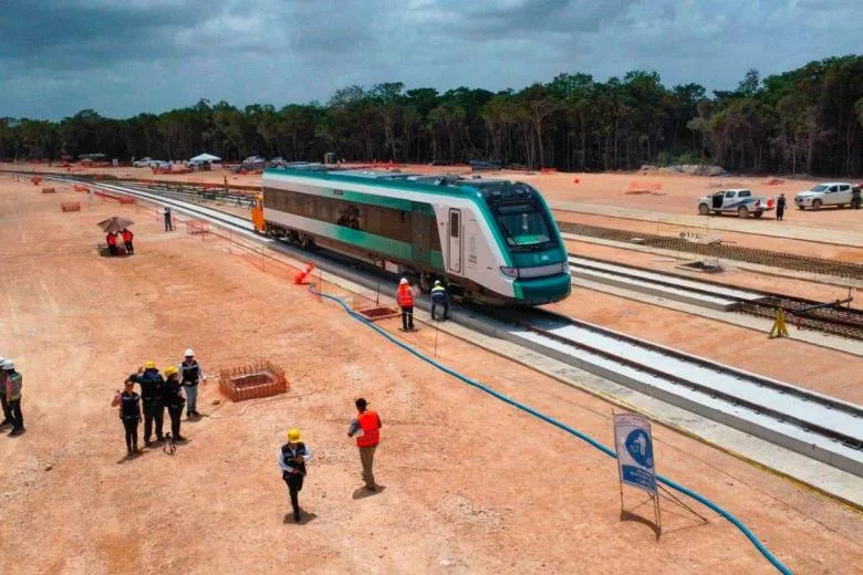 A newly constructed train on railway tracks surrounded by construction workers in a wooded area.