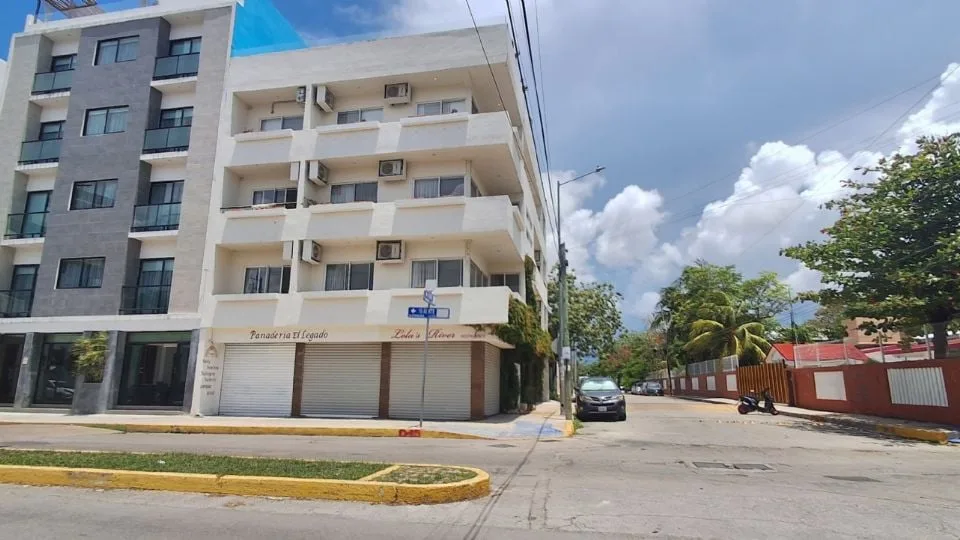 View of a modern multi-story building with balconies and storefronts, surrounded by trees on a sunny day