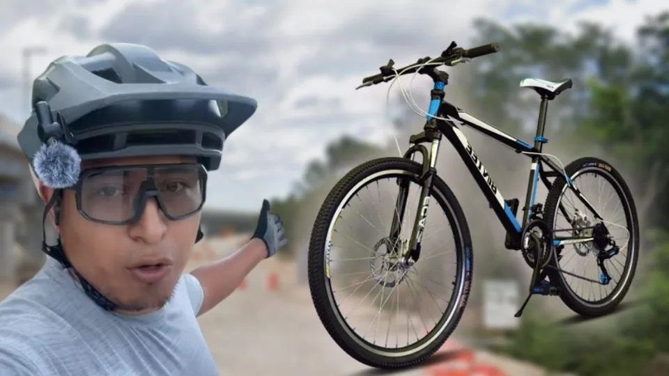 A person wearing a helmet and glasses stands next to a mountain bike while gesturing towards a path in the background.$# CAPTION