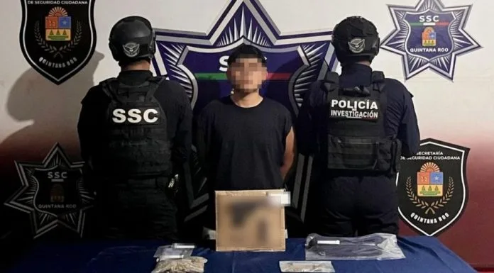 A suspect is flanked by two police officers wearing tactical gear, in a setting with police insignia and a table displaying evidence related to the arrest.$# CAPTION