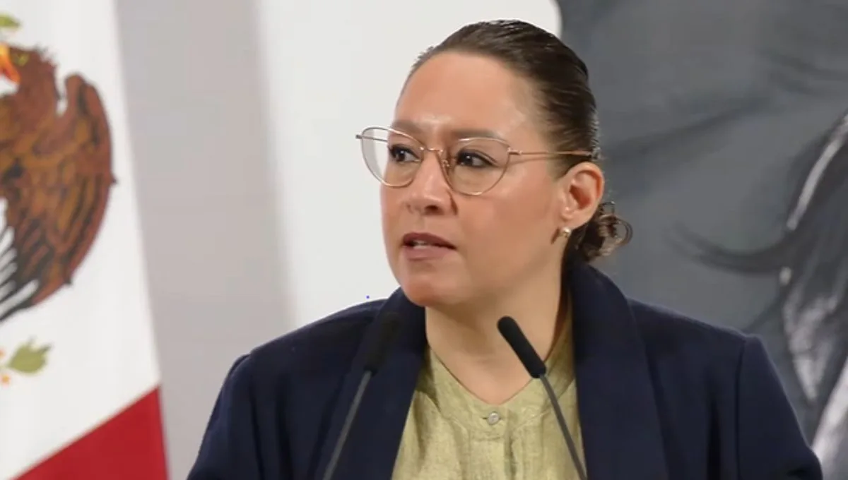 A woman with glasses speaking at a podium, with a Mexican flag in the background