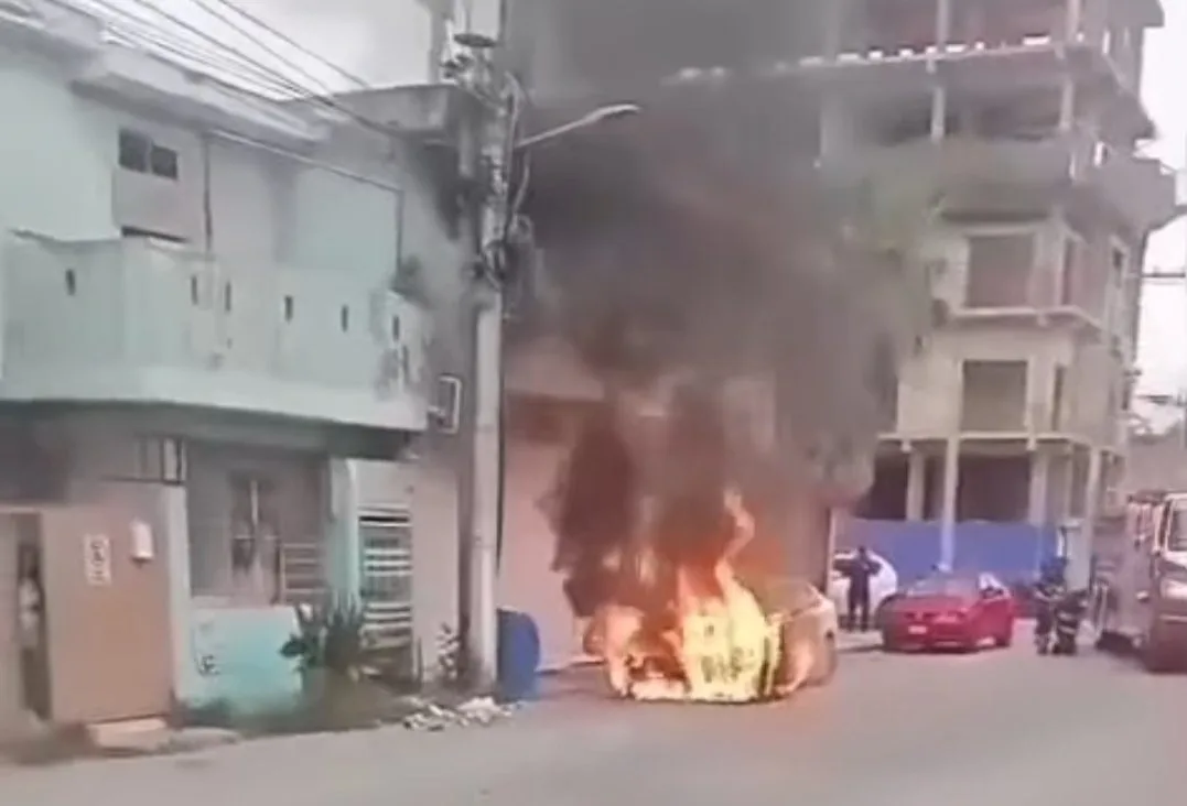 A car engulfed in flames on a city street, with smoke billowing into the air and nearby buildings visible in the background.$# CAPTION
