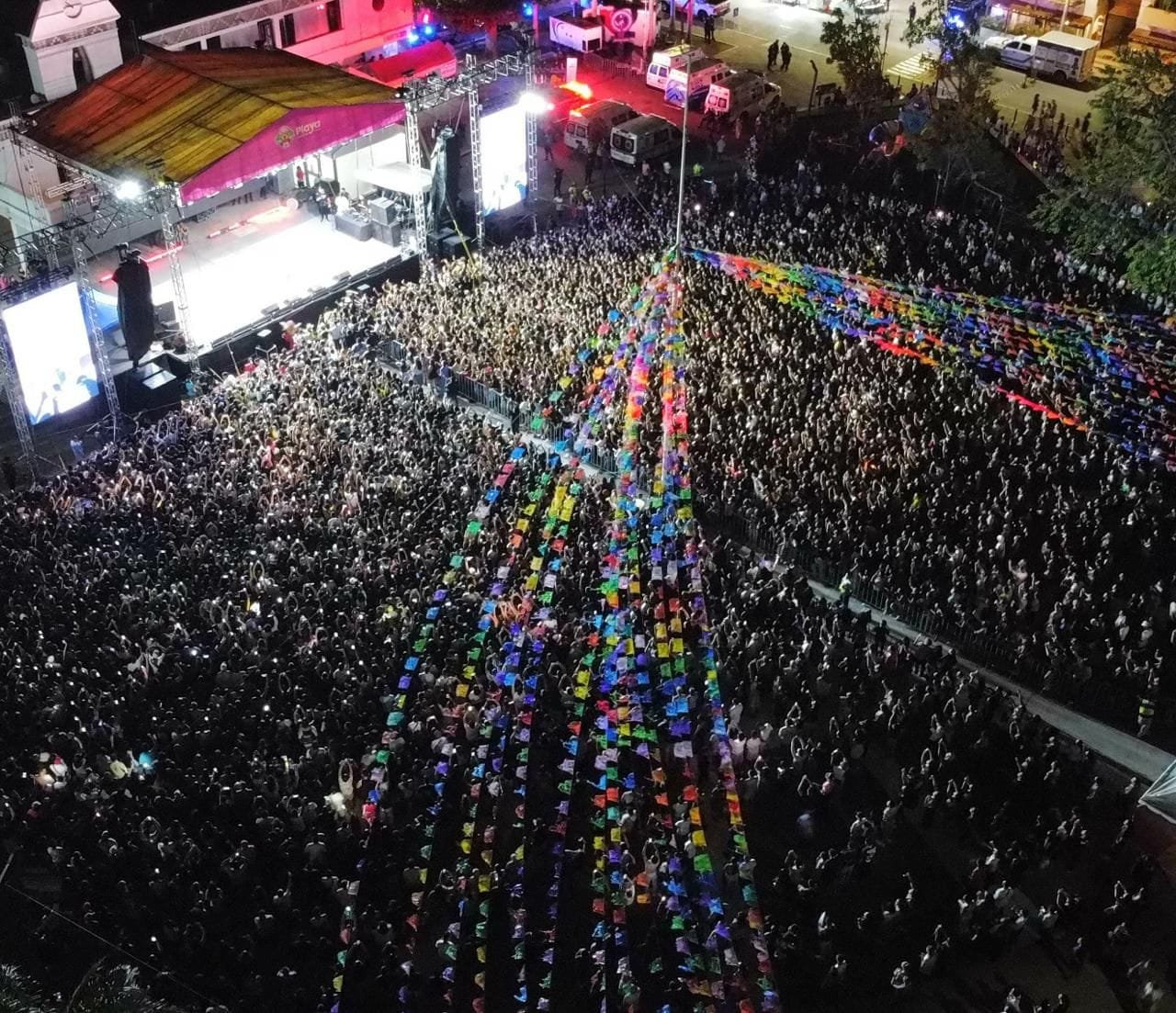 Aerial view of a large crowd at a concert with colorful decorations and lights in the background.$# CAPTION