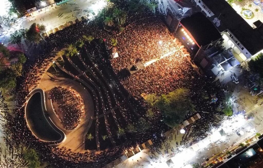 Aerial perspective of a massive crowd gathered at night for an outdoor event, with a stage illuminated in the background and surrounding trees and structures visible.