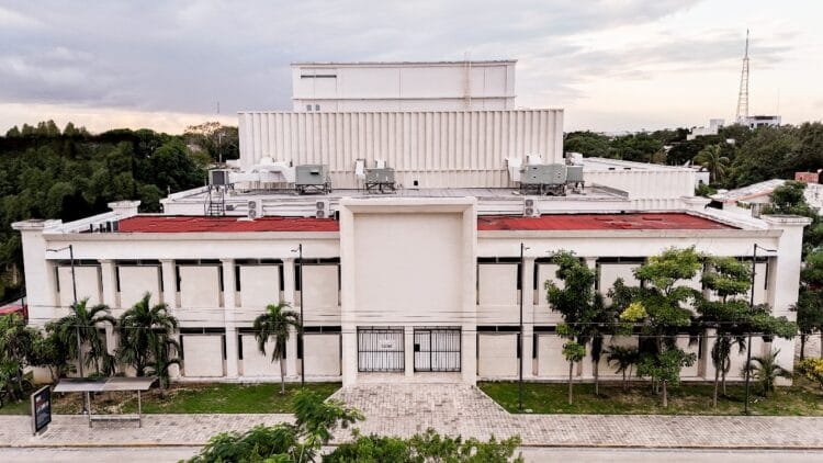 An aerial view of a modern building with a flat roof, surrounded by trees and greenery, under a cloudy sky