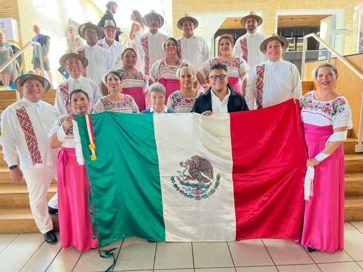 A group of people dressed in traditional Mexican attire holding a large Mexican flag, smiling, and posing for a photo indoors.$#$ CAPTION