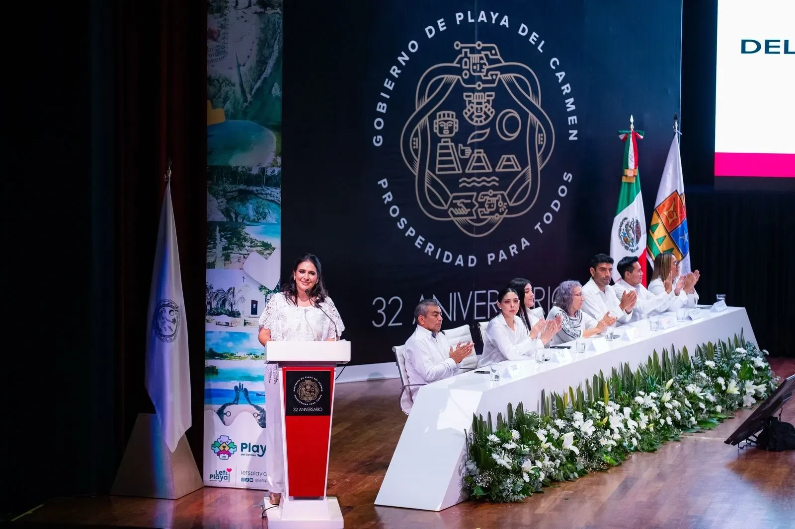 A woman addresses the audience at the 32nd anniversary event of Playa del Carmen, surrounded by seated attendees on a stage decorated with greenery and flags.$# CAPTION