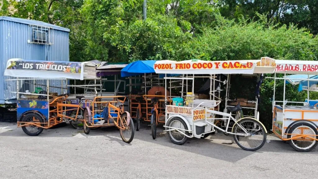 A row of colorful food carts, including three tricycles for selling snacks and drinks, parked in a green area.$# CAPTION