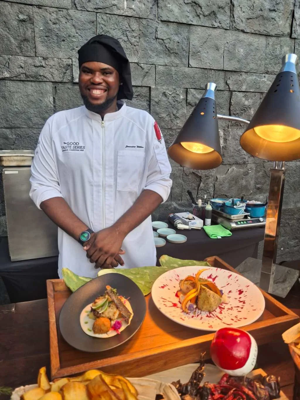 A smiling chef stands proudly next to beautifully plated dishes at a culinary event, with stone walls in the background.$# CAPTION
