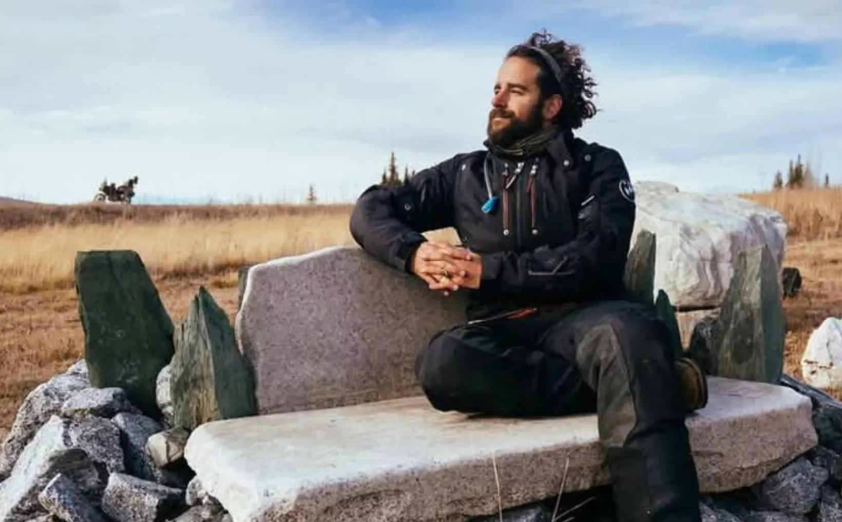 A person sitting on a stone bench surrounded by grass and rocky terrain under a blue sky.