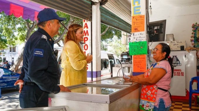 A police officer and a woman engage in conversation at a food stall, with a smiling vendor serving ice cream in a colorful market setting.$#$ CAPTION