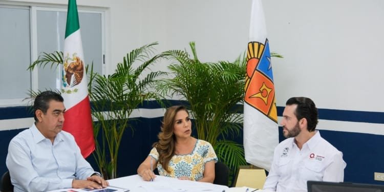 Three people discussing in a government office setting, with flags in the background and papers on the table.