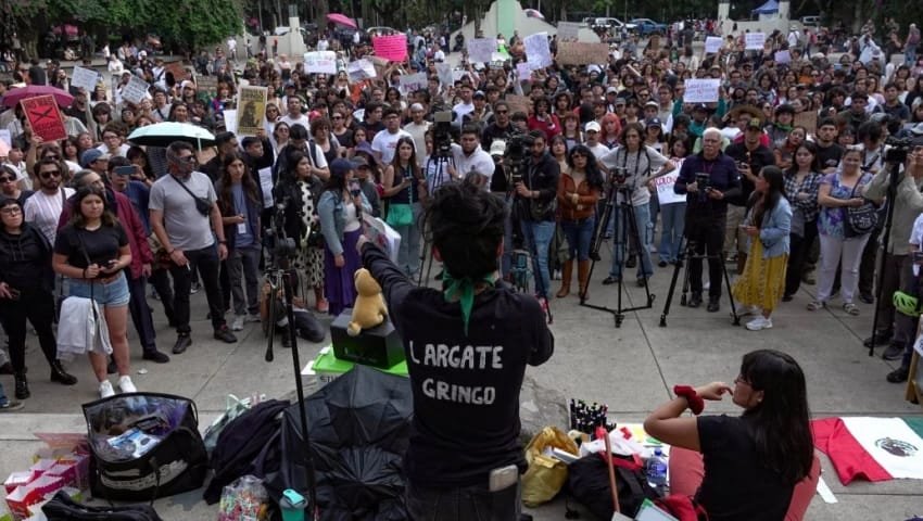 A crowd of people at a protest holding signs, with a speaker visible in the foreground. The speaker has their back to the camera and is wearing a black shirt with the text "LARGATE GRINGO."